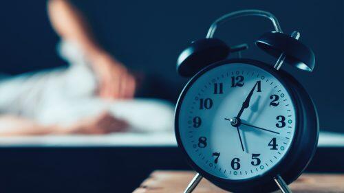 Sleeping disorder or insomnia concept, selective focus of vintage clock in bedroom and out of focus male person trying to fall asleep in bed